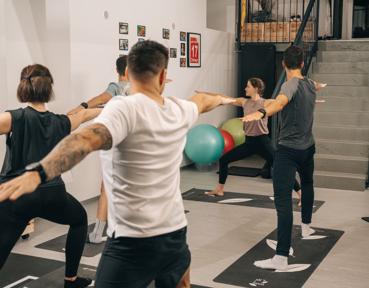 Gruppen-Yoga Training von Trainerin Chrisi mit Teilnehmern in der stehenden Yoga-Pose Krieger 2, Sicht von hinten