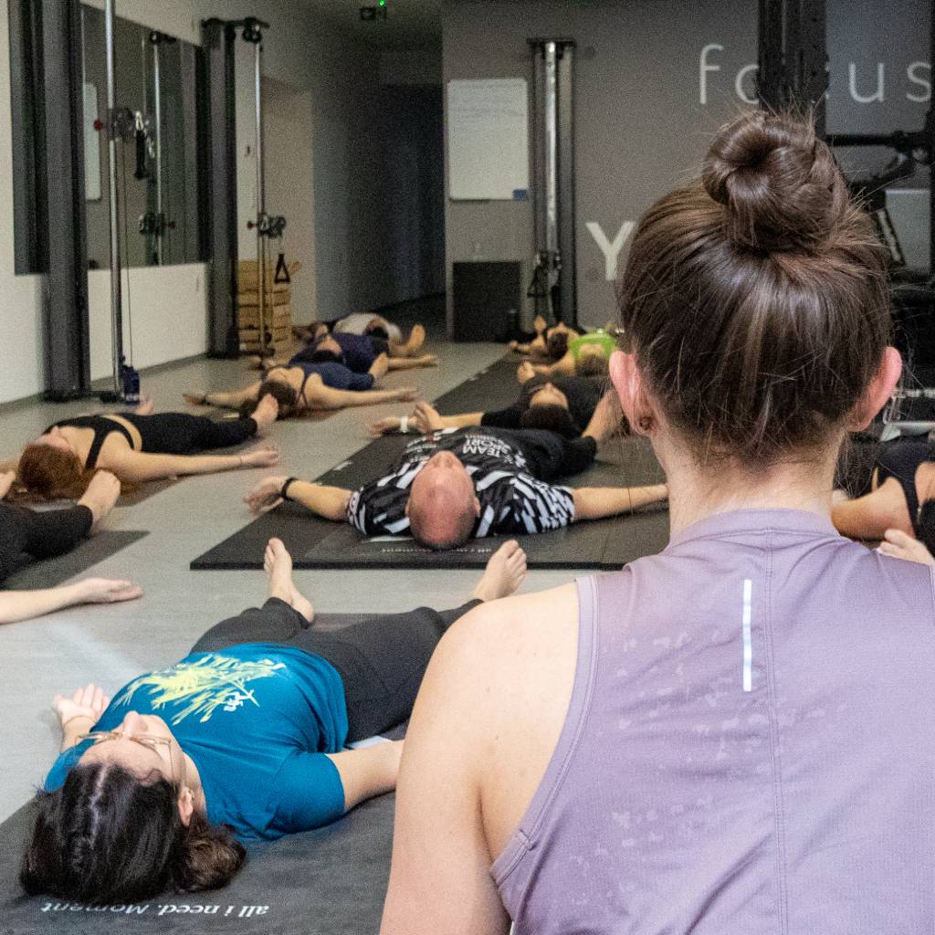 Yoga-Trainerin Chrisi sitzend vor den Teilnehmer:innen in der liegenden Schlussentspannung